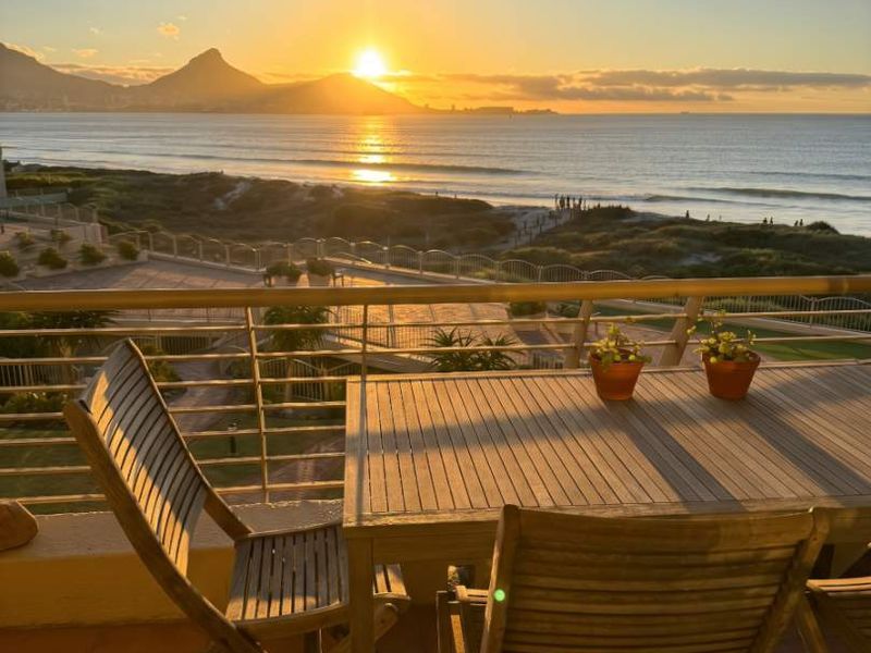 A spacious balcony featuring tiled flooring and a metal balustrade, offering stunning sunset views over the ocean and coastline.