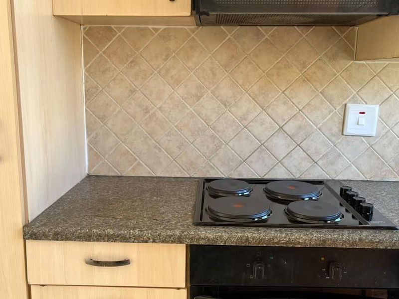 A functional kitchen area featuring light wood-finish cabinetry, a dark laminate countertop, a built-in electric hob, oven, and a neutral tiled backsplash.