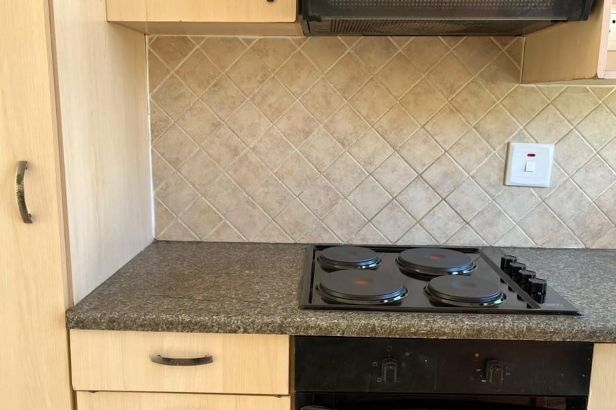 A functional kitchen area featuring light wood-finish cabinetry, a dark laminate countertop, a built-in electric hob, oven, and a neutral tiled backsplash.