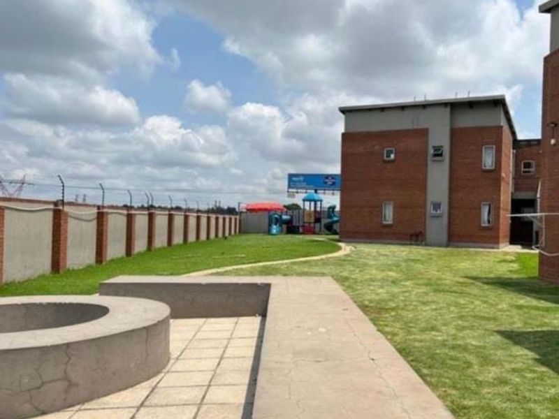 Rear exterior view of the apartment complex featuring a communal lawn, paved patio area with a built-in concrete fire pit, and perimeter security fencing.