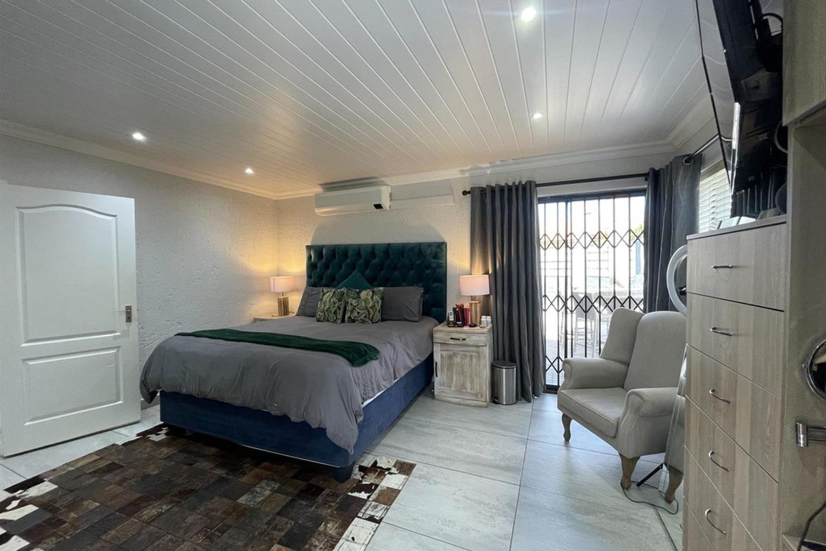 A well-maintained bedroom featuring large floor tiles, a tongue-and-groove ceiling with recessed lighting, a wall-mounted air conditioning unit, and sliding glass doors with security gates.