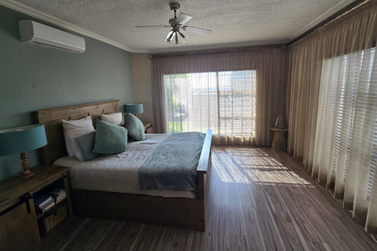 Spacious bedroom featuring wood-look flooring, a wall-mounted air conditioning unit, ceiling fan, and large windows with floor-to-ceiling curtains.
