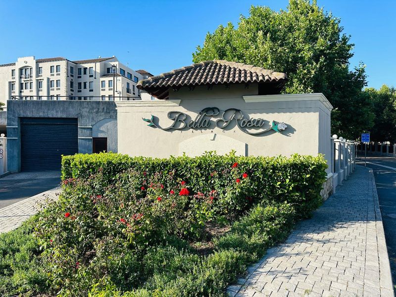 The entrance to Bella Rosa Village, featuring a well-maintained complex sign, landscaped garden beds, and paved access.