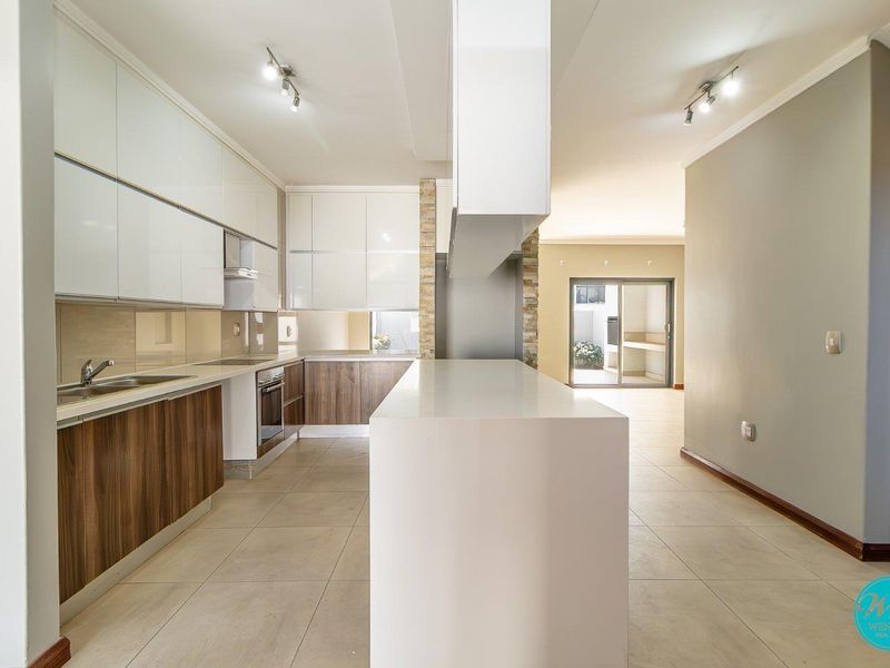 Modern kitchen featuring high-gloss cabinetry, a central island, integrated appliances, and neutral tiled flooring, leading to an open-plan living area.