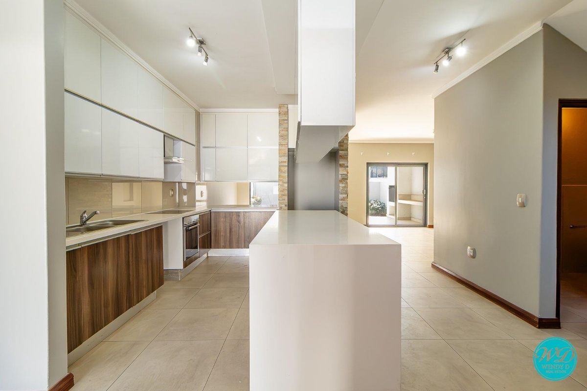 Modern kitchen featuring high-gloss cabinetry, a central island, integrated appliances, and neutral tiled flooring, leading to an open-plan living area.