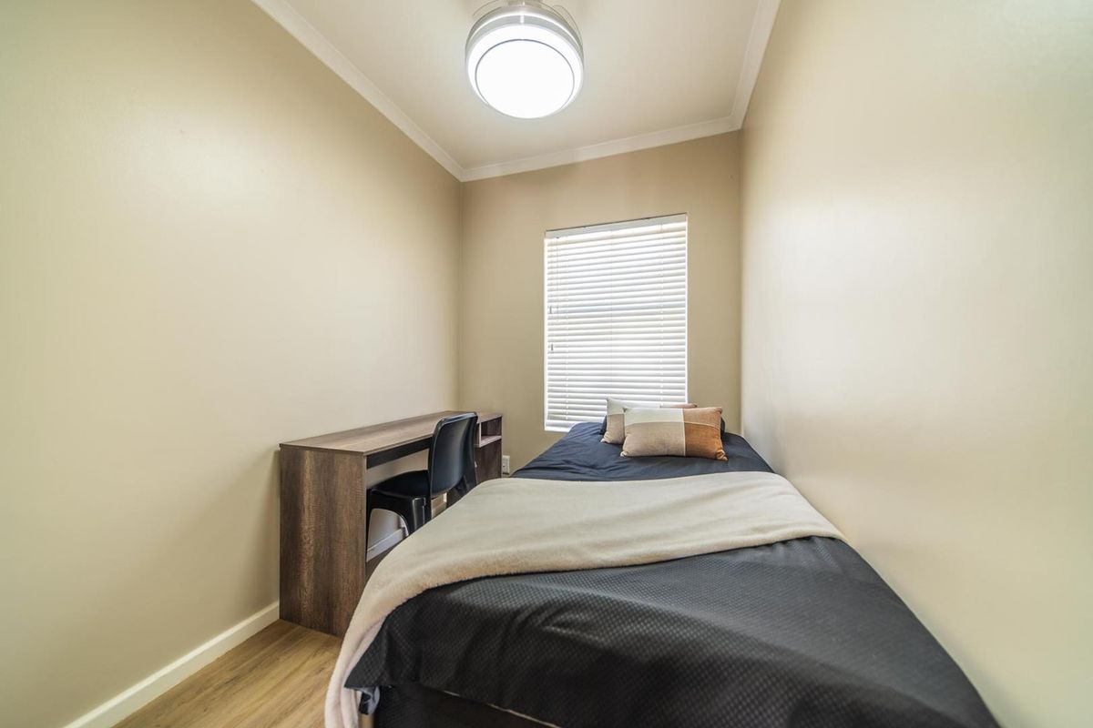 A compact bedroom featuring neutral wall tones, wood-look flooring, a central ceiling light fixture, and a window fitted with horizontal blinds.