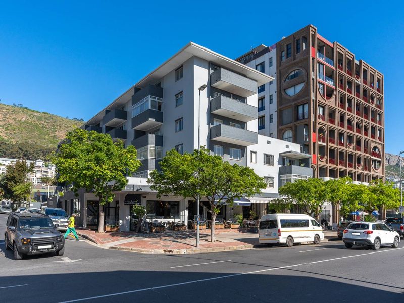 A street-level view of a modern apartment building featuring balconies and ground-floor commercial space in a vibrant urban setting.
