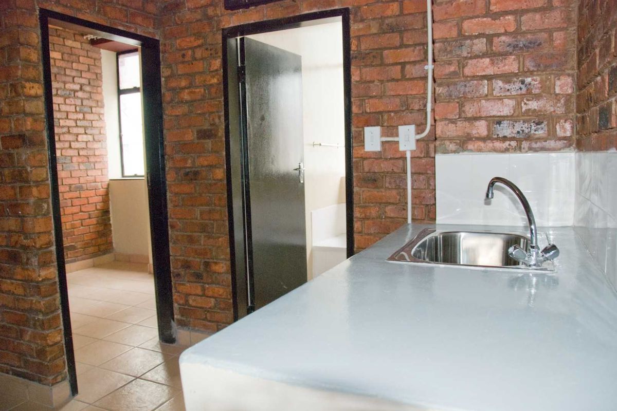 A compact kitchen area featuring exposed brick walls, a stainless steel sink, and a painted concrete countertop, leading into adjacent rooms.