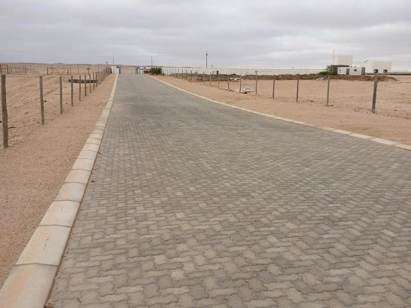 A view of a paved residential driveway leading into a vacant land development, featuring concrete kerbing and wire perimeter fencing.