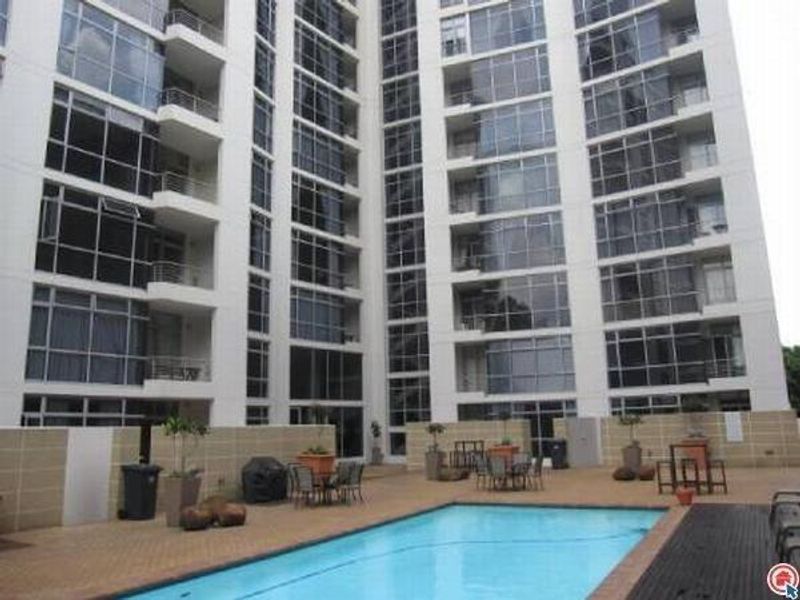 A communal swimming pool area set against a modern high-rise apartment building with extensive glass facade and private balconies.