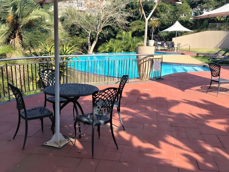 A well-maintained outdoor pool area featuring a paved terrace, metal safety fencing, and a fixed shade umbrella for communal relaxation.