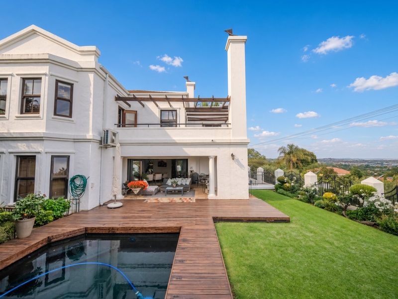 Rear elevation featuring a swimming pool with timber decking, a covered patio, an upper-level balcony, and a well-maintained garden with scenic views.