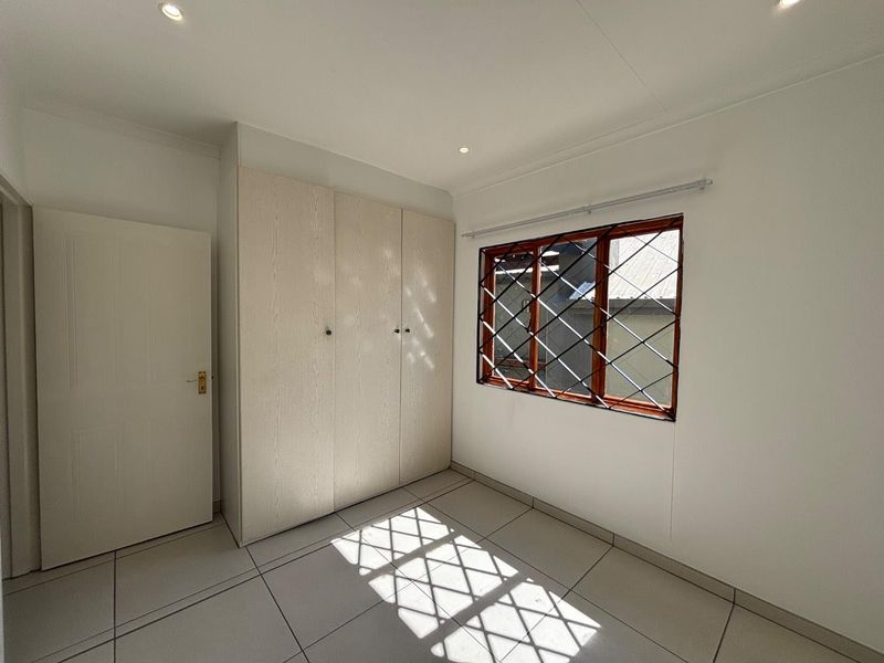 A bright, empty bedroom featuring neutral tiled flooring, built-in cupboards, and a window fitted with security bars.