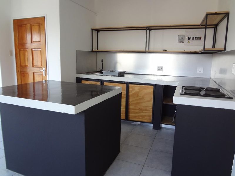 A modern industrial-style kitchen featuring a central island, stainless steel splashback, built-in hob, and open shelving, currently in an unfinished state.