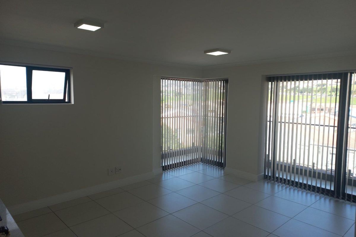 A bright, empty living area featuring neutral tiled flooring, modern recessed lighting, and large windows with vertical blinds providing ample natural light.