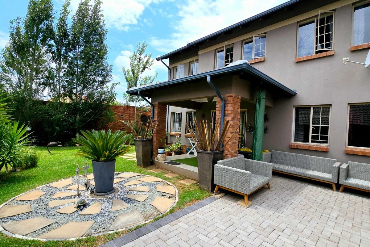Rear view of the townhouse featuring a covered patio with brick pillars, paved courtyard, and a landscaped garden with a circular stone feature.
