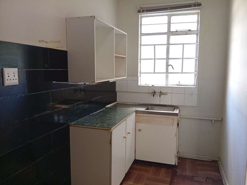 A compact, dated kitchen featuring basic built-in cabinetry, a stainless steel sink, and dark tiled splashbacks, requiring modernization and cosmetic updates.