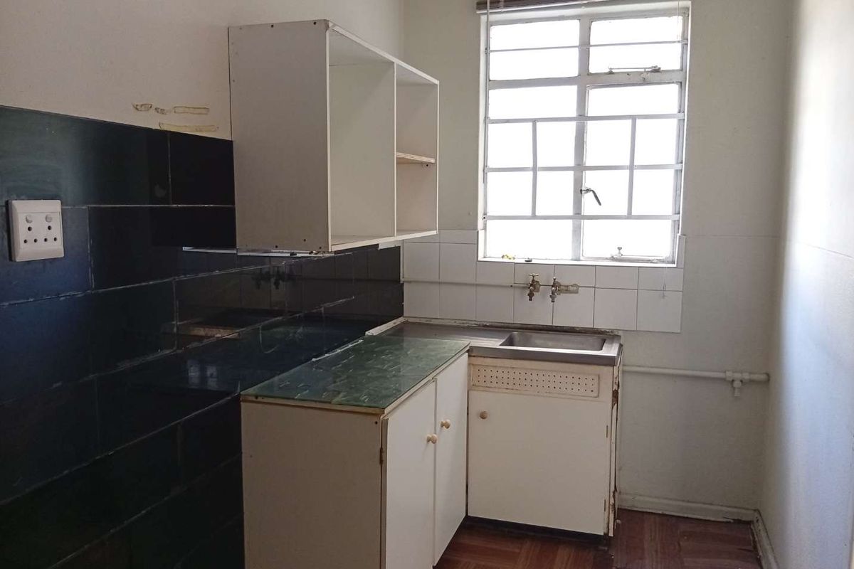 A compact, dated kitchen featuring basic built-in cabinetry, a stainless steel sink, and dark tiled splashbacks, requiring modernization and cosmetic updates.