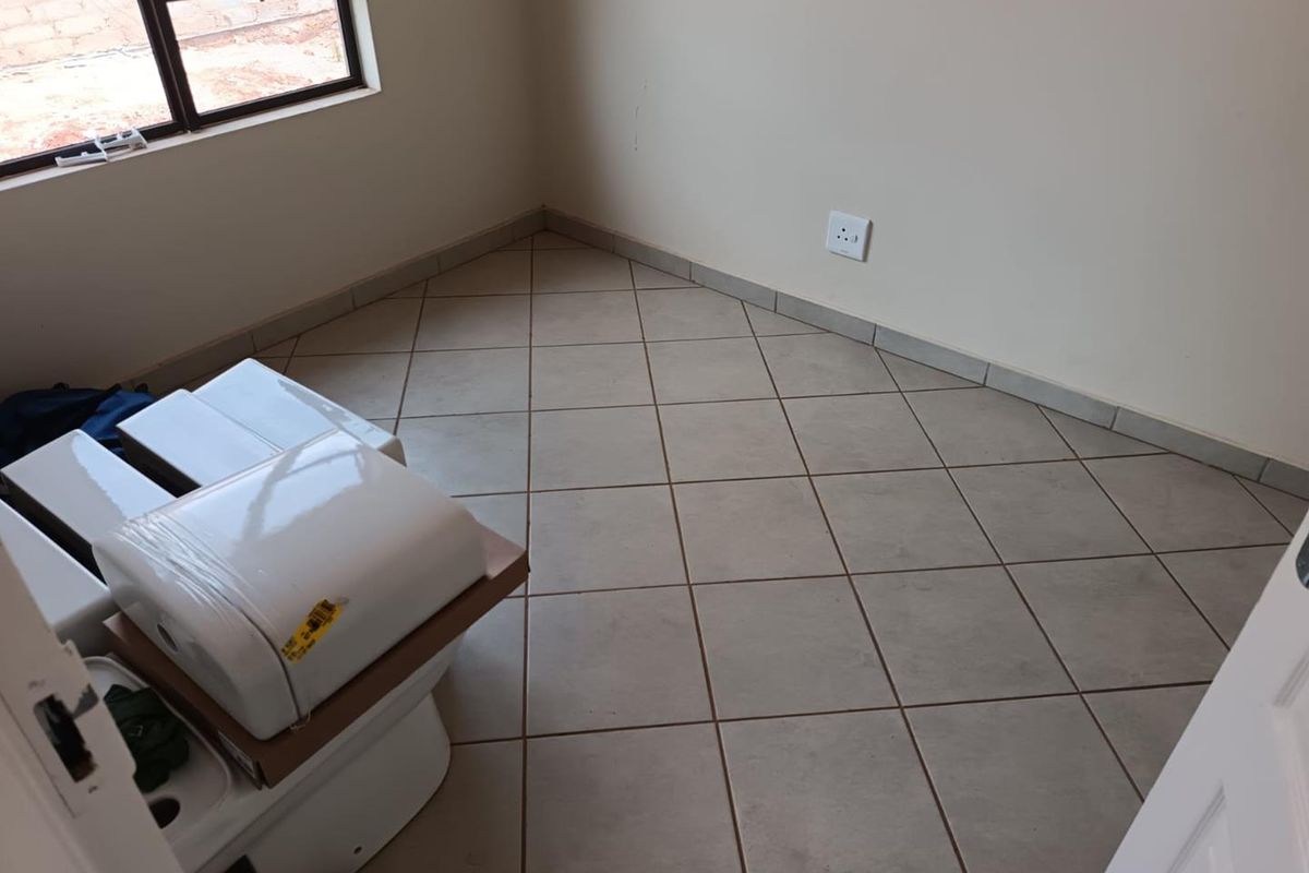A small, empty room featuring tiled flooring and a window, currently used for storing boxed bathroom fixtures.