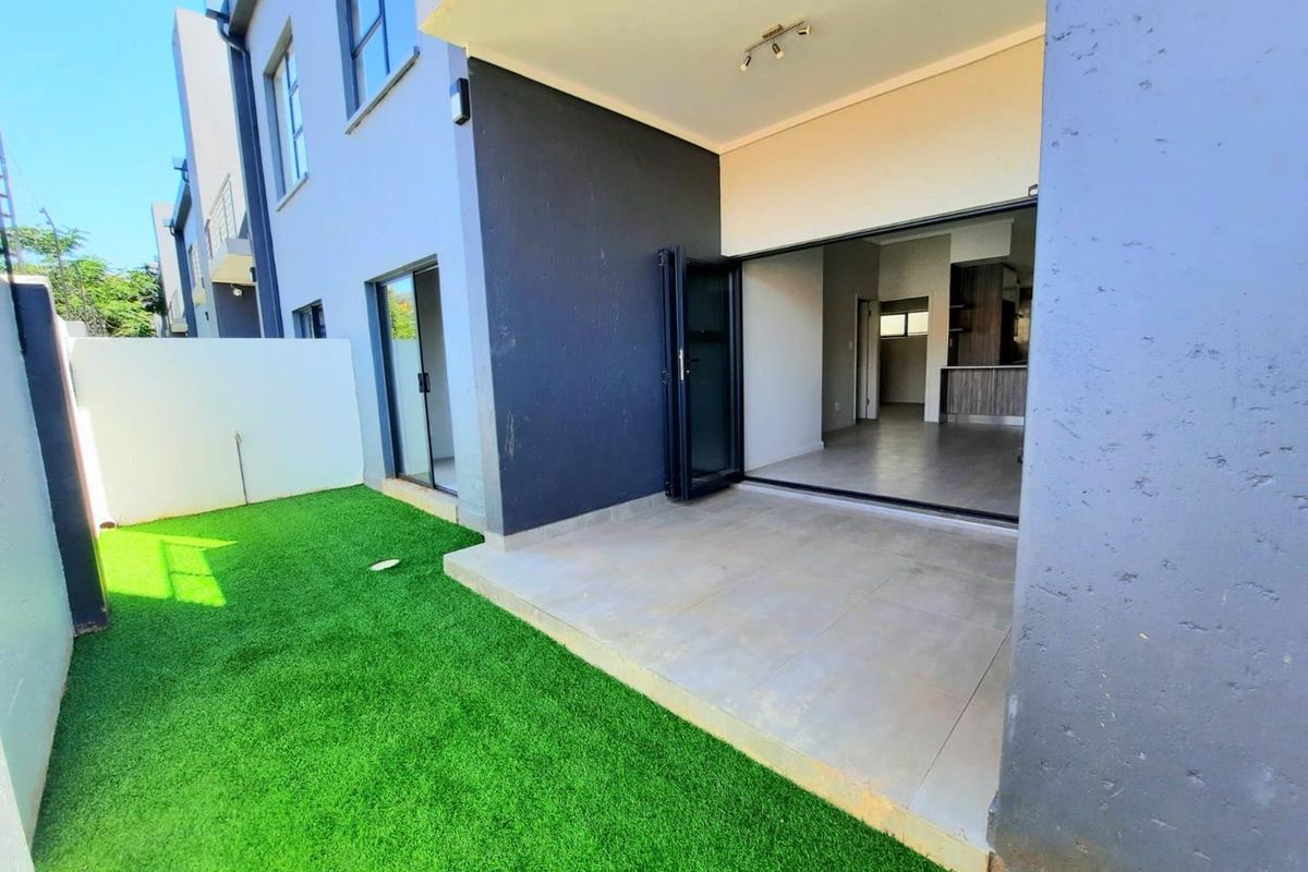 A modern rear exterior view featuring a covered patio with tiled flooring, artificial grass lawn, and seamless indoor-outdoor flow through large sliding and bi-fold doors.