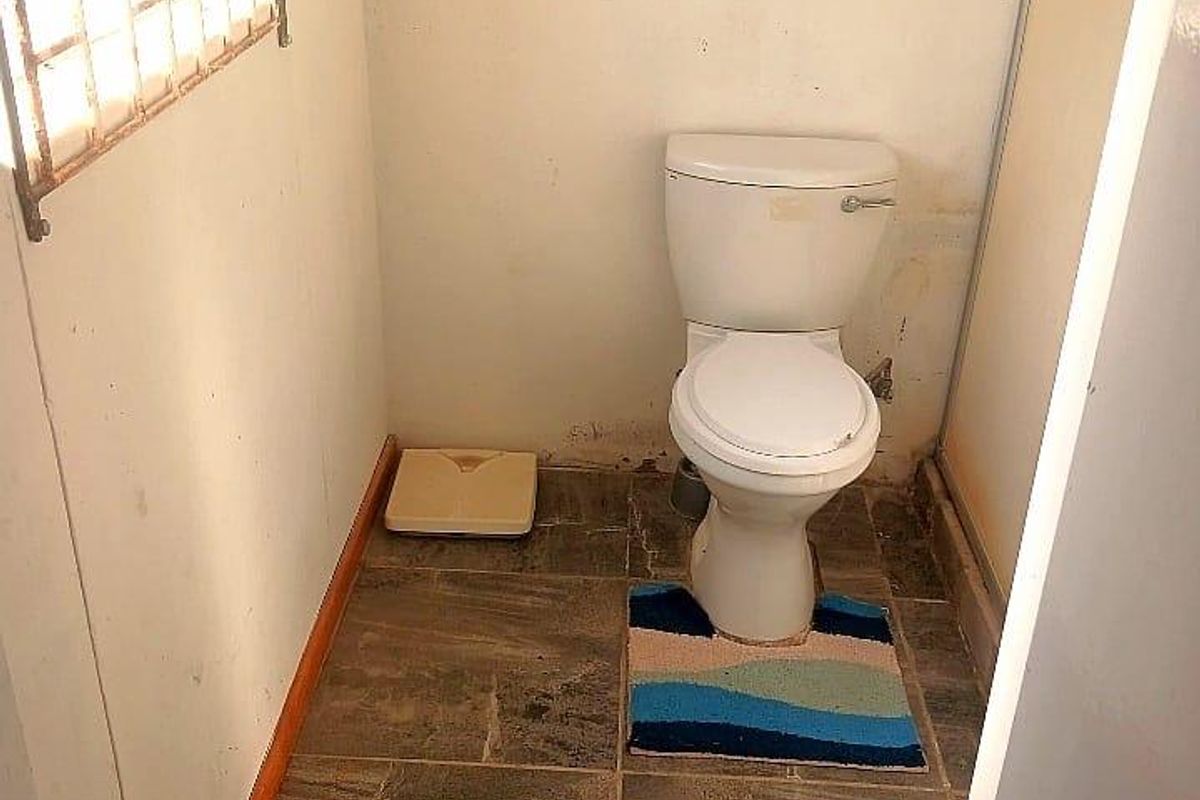A small, functional bathroom featuring tiled flooring, a standard toilet, and a window with security bars, requiring cosmetic updates to improve its condition.