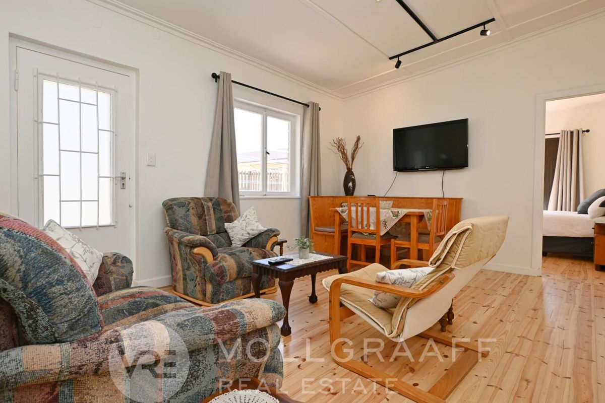 A living room featuring light-toned timber flooring, crown molding, and track lighting, with a doorway leading to an adjacent bedroom.