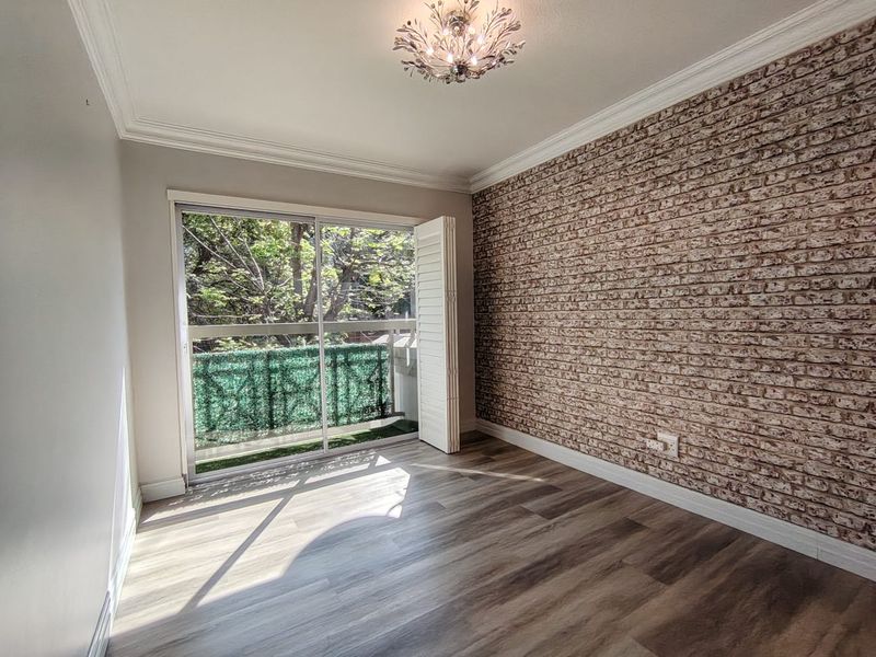 A bright, empty bedroom featuring modern laminate flooring, a decorative faux brick accent wall, and sliding glass doors opening onto a private balcony.