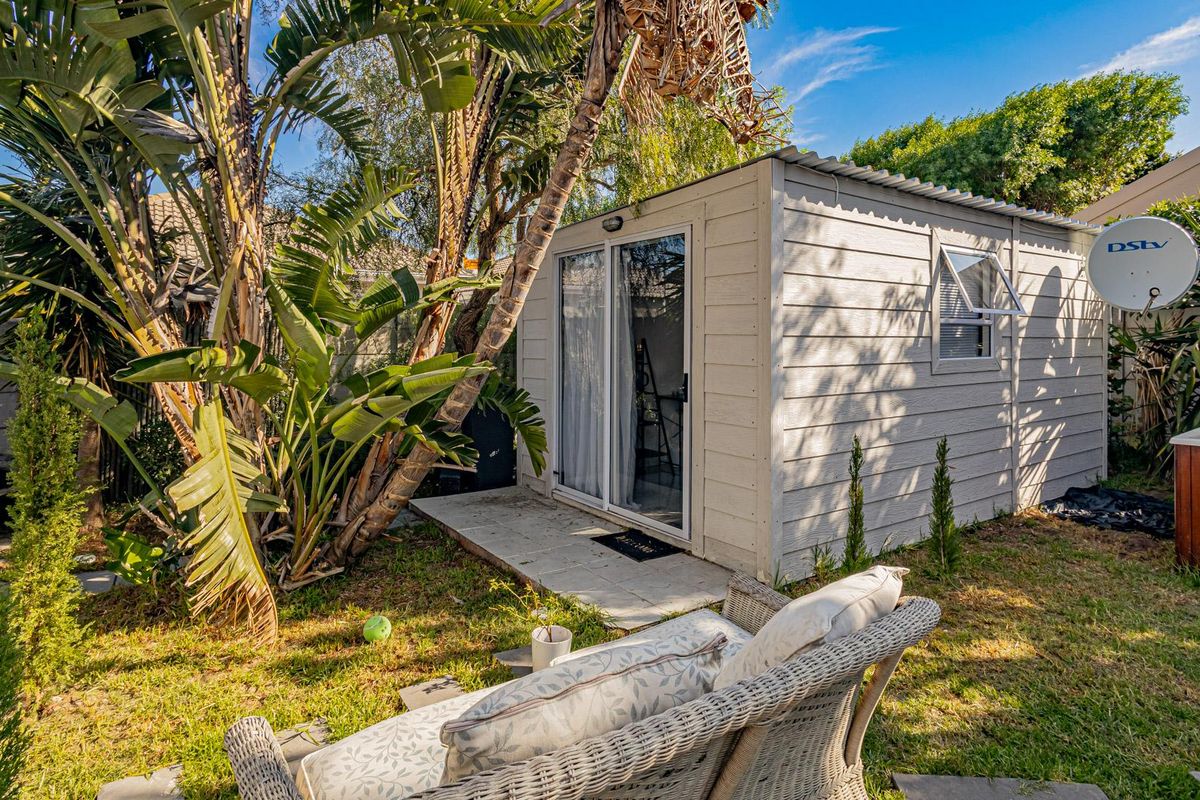 A detached garden studio or utility structure featuring a sliding glass door, corrugated roof, and paved patio area set within a landscaped garden.