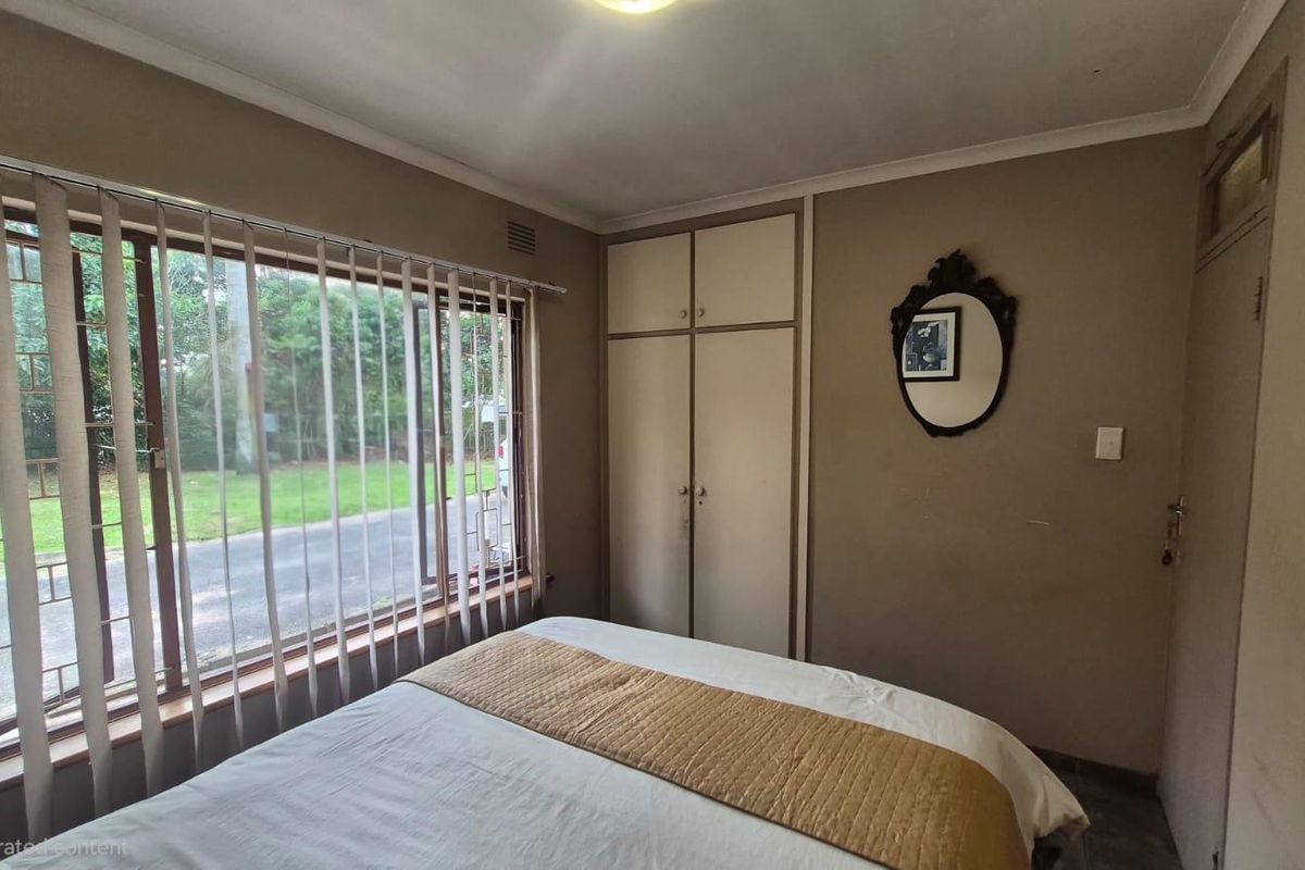 A functional bedroom featuring built-in cupboards, large windows with vertical blinds, and neutral wall tones, offering a comfortable and practical living space.