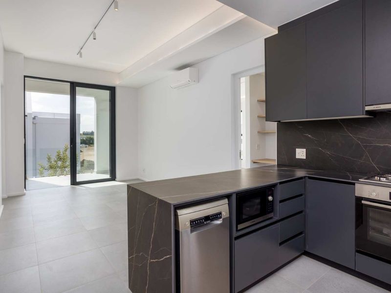 Modern open-plan kitchen and living area featuring sleek dark cabinetry, stone countertops, integrated appliances, and direct access to a private balcony.