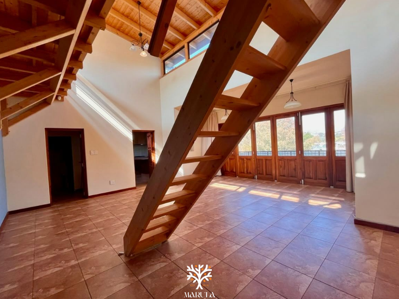Spacious open-plan living area featuring high exposed timber ceilings, a central wooden staircase, tiled floors, and large timber-framed glass doors leading to the exterior.