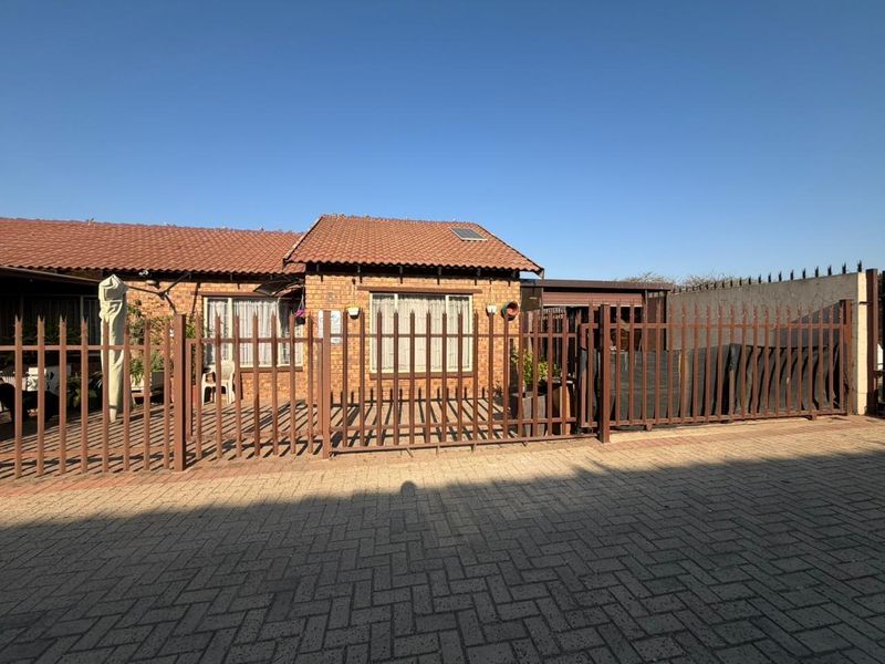 Front view of a brick house featuring a tiled roof, paved driveway, and secure metal perimeter fencing.