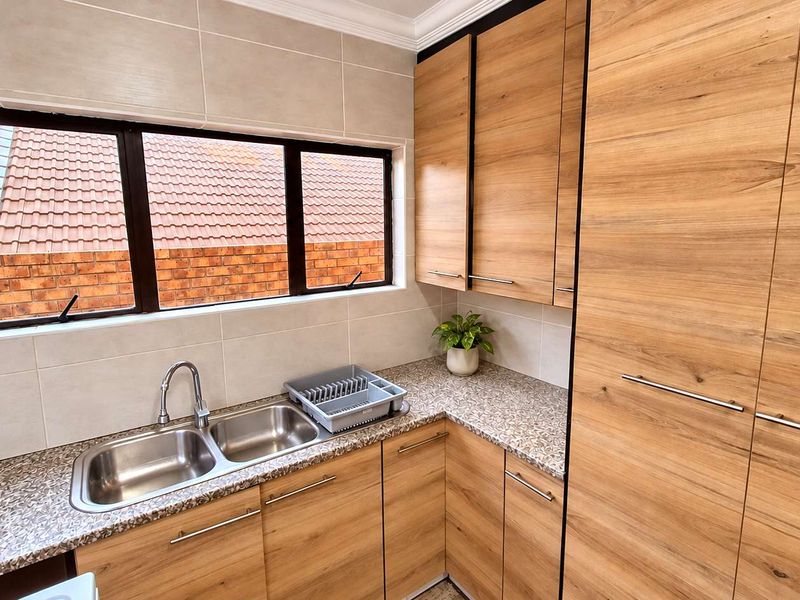 A modern, well-maintained scullery featuring wood-finish cabinetry, granite countertops, and a double stainless steel sink with a view of the exterior.