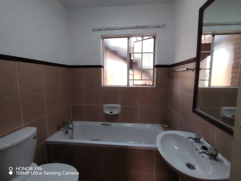 A functional bathroom featuring a bathtub, pedestal basin, and toilet, finished with mid-tone wall tiles and a window with security bars.