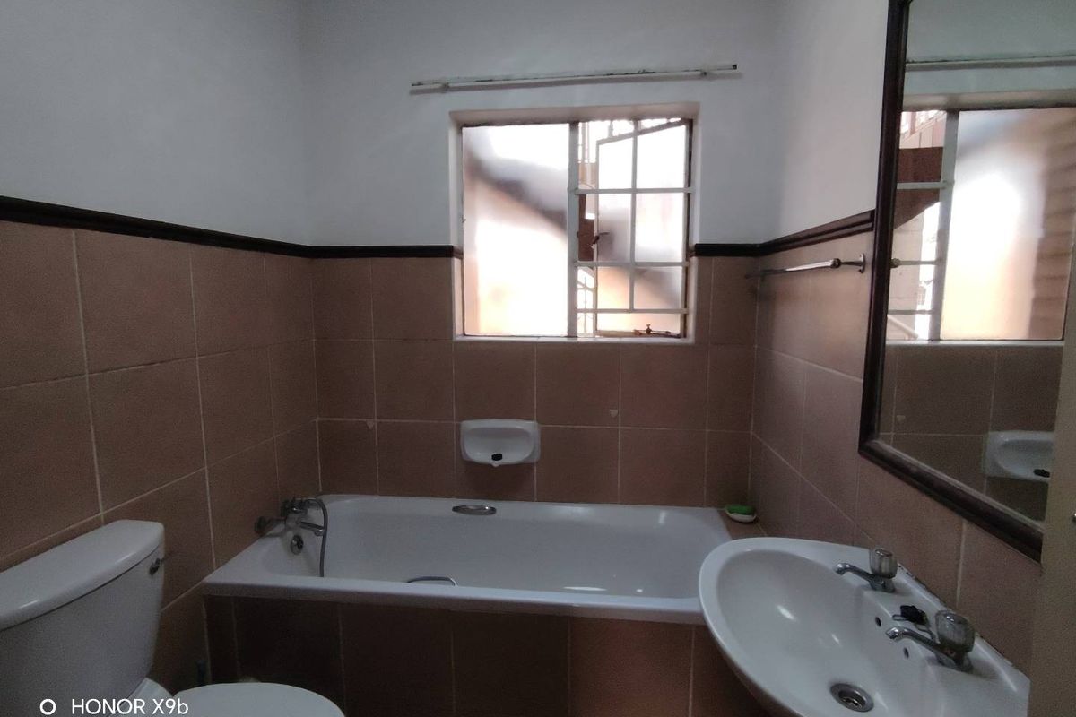 A functional bathroom featuring a bathtub, pedestal basin, and toilet, finished with mid-tone wall tiles and a window with security bars.