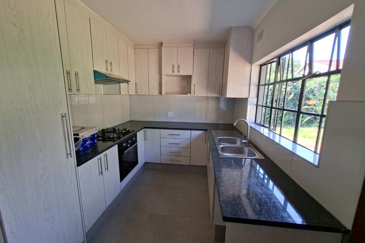 A clean, modern kitchen featuring light wood-grain cabinetry, dark granite countertops, a gas hob, built-in oven, and a double stainless steel sink.