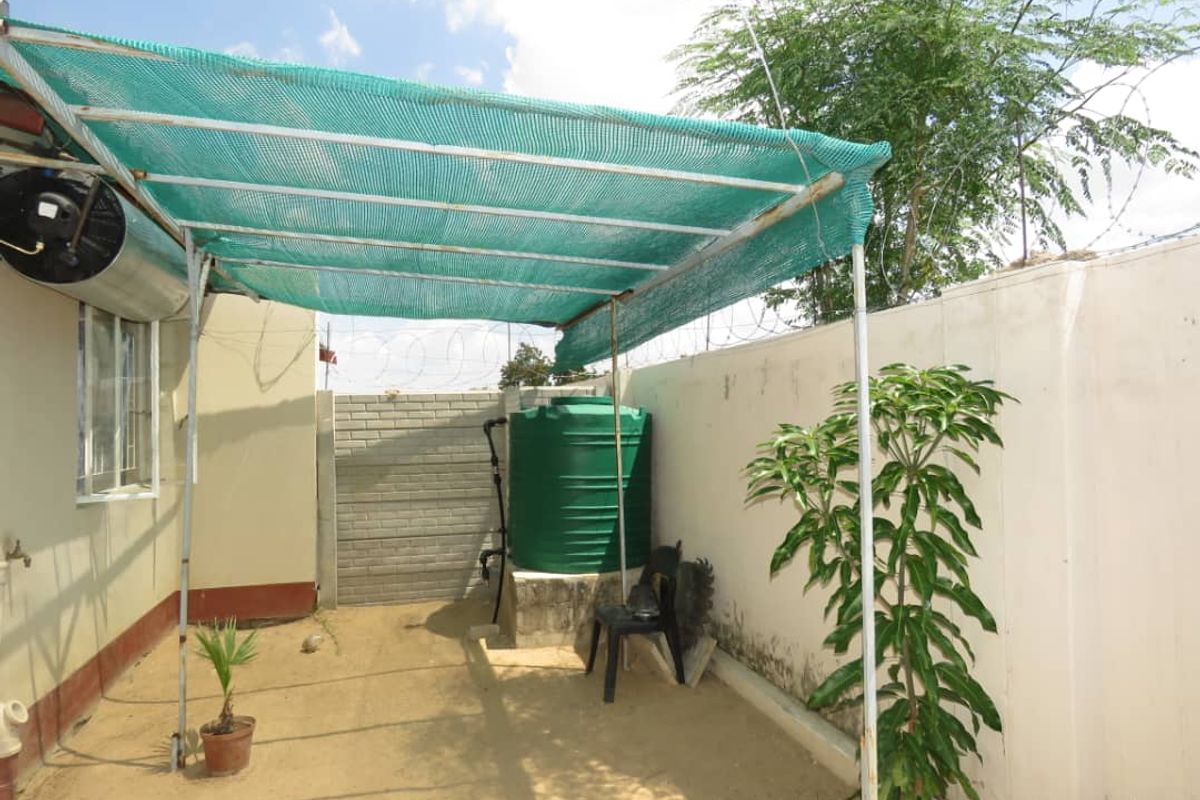 An outdoor utility area featuring a shade cloth canopy, a water storage tank, and perimeter walling with security fencing.