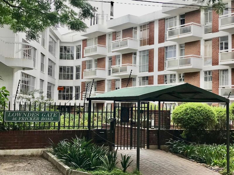 Exterior view of Lowndes Gate apartment complex featuring a brick facade, private balconies, secure gated entrance, and established garden landscaping.