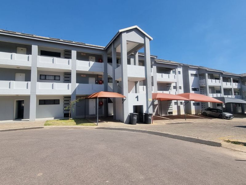 Exterior view of a multi-story apartment complex featuring paved driveway access, covered parking bays, and a central stairwell entrance.