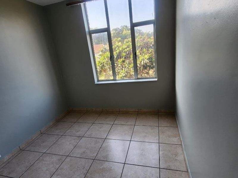 An empty bedroom featuring tiled flooring, neutral wall paint, and a large window providing natural light and a view of the surrounding greenery.