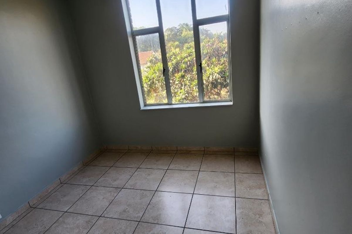 An empty bedroom featuring tiled flooring, neutral wall paint, and a large window providing natural light and a view of the surrounding greenery.