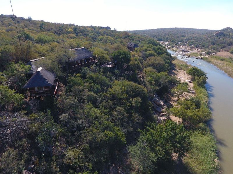 Aerial view of a bushveld lodge featuring multiple thatched-roof units situated on a riverbank, surrounded by natural vegetation.