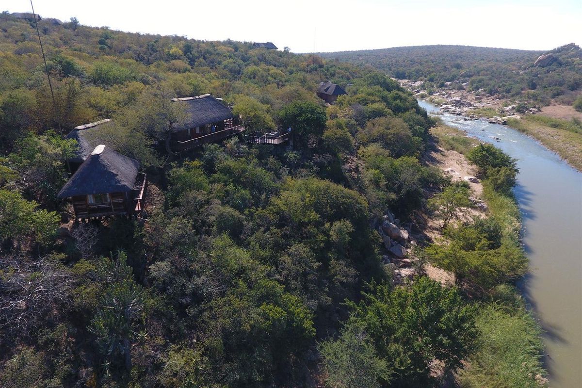 Aerial view of a bushveld lodge featuring multiple thatched-roof units situated on a riverbank, surrounded by natural vegetation.