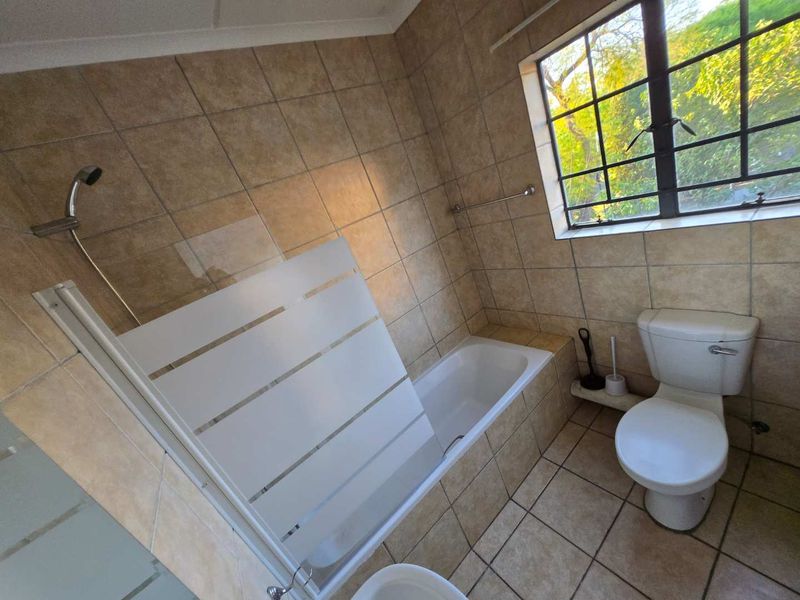 A functional bathroom featuring a built-in bathtub with a shower screen, a toilet, and neutral floor-to-ceiling tiling.