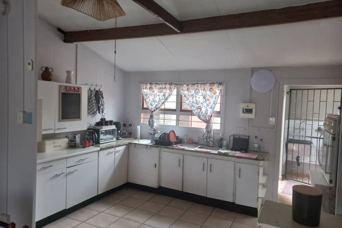A functional kitchen featuring white cabinetry, laminate countertops, double sink, and exposed timber ceiling beams, with tiled flooring throughout.