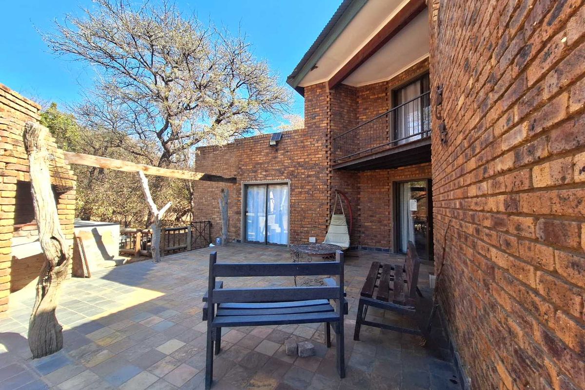 Rear exterior view featuring a spacious tiled patio, exposed brick facade, built-in braai area, and an upper-level balcony with metal railings.