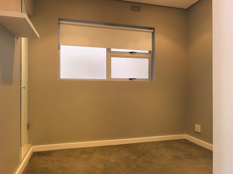 A clean, neutral-toned bedroom featuring carpeted flooring, a built-in cupboard, and a window with a roller blind.