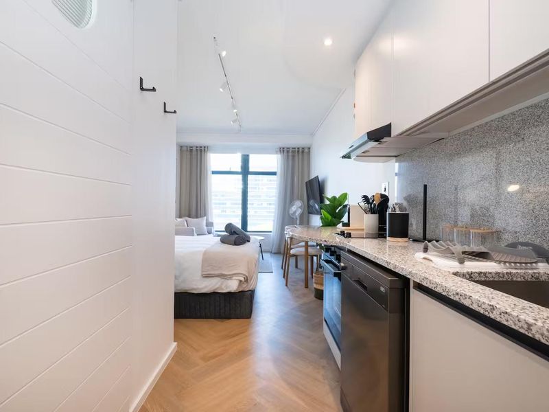 A modern open-plan studio apartment featuring a compact kitchen with granite countertops, built-in appliances, and elegant herringbone wood flooring throughout.