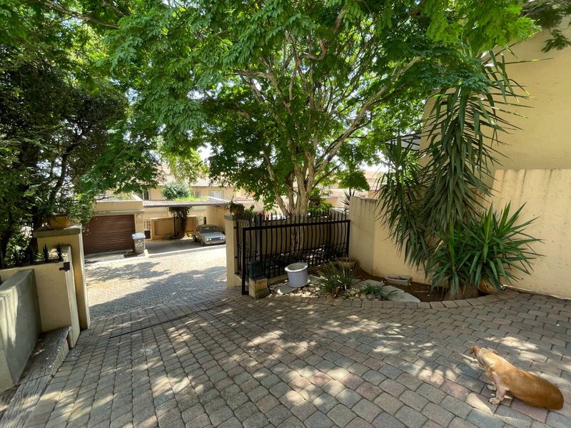 A paved driveway entrance to the townhouse property featuring mature trees, a metal security gate, and a retaining wall.