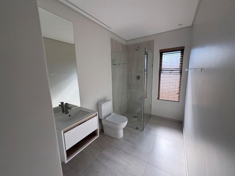 Modern bathroom featuring a sleek vanity, glass-enclosed shower, neutral floor tiles, and a window with fitted blinds, presenting a clean and contemporary aesthetic.